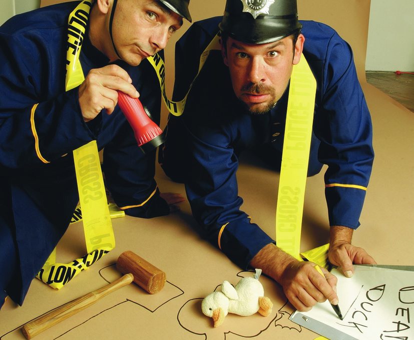 Two actors dressed as police officers humorously investigate a mock crime scene in a playful setup for a Murder Mystery Dinner event. They are kneeling on the ground, surrounded by yellow police line tape, with a chalk outline on the floor resembling the shape of a duck. One officer is holding a red flashlight while the other points at a toy duck with a sign labeled 'Dead Duck.' The scene is lighthearted and comedic, emphasizing the fun, interactive nature of the mystery event.