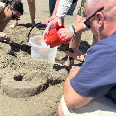 Team members collaborating on their sand sculpture, using buckets and tools to shape the design during a sand sculpting team building event at the beach. thumbnail