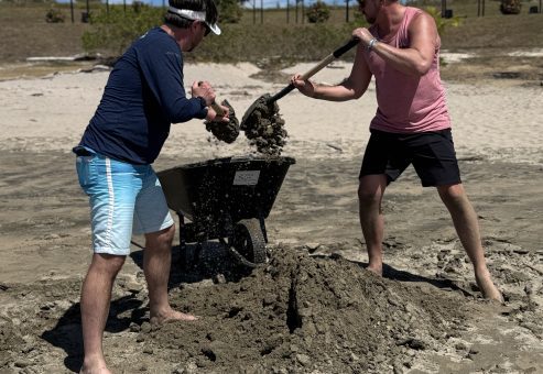 Two team members collaborating to transport sand with a wheelbarrow during a sand sculpting team building event.