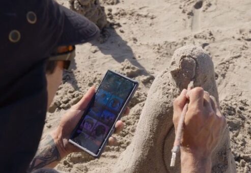 A participant refining their sand sculpture while using a reference image on their phone during a sand sculpting team building event.
