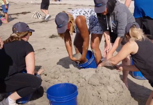 Team collaborating on shaping a sand sculpture using buckets of sand at a sand sculpting team building event on the beach.
