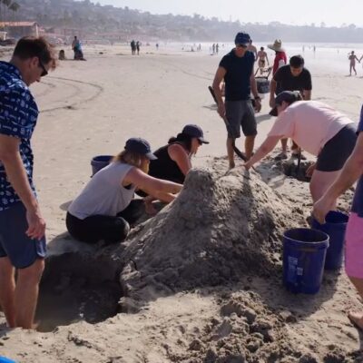 A team working together at the beach to build a large sand sculpture during a sand sculpting team building event, with teamwork and coordination. thumbnail