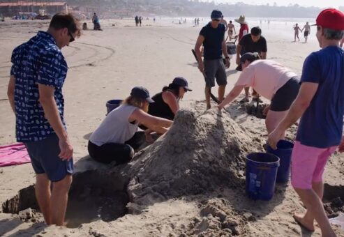 A team working together at the beach to build a large sand sculpture during a sand sculpting team building event, with teamwork and coordination.