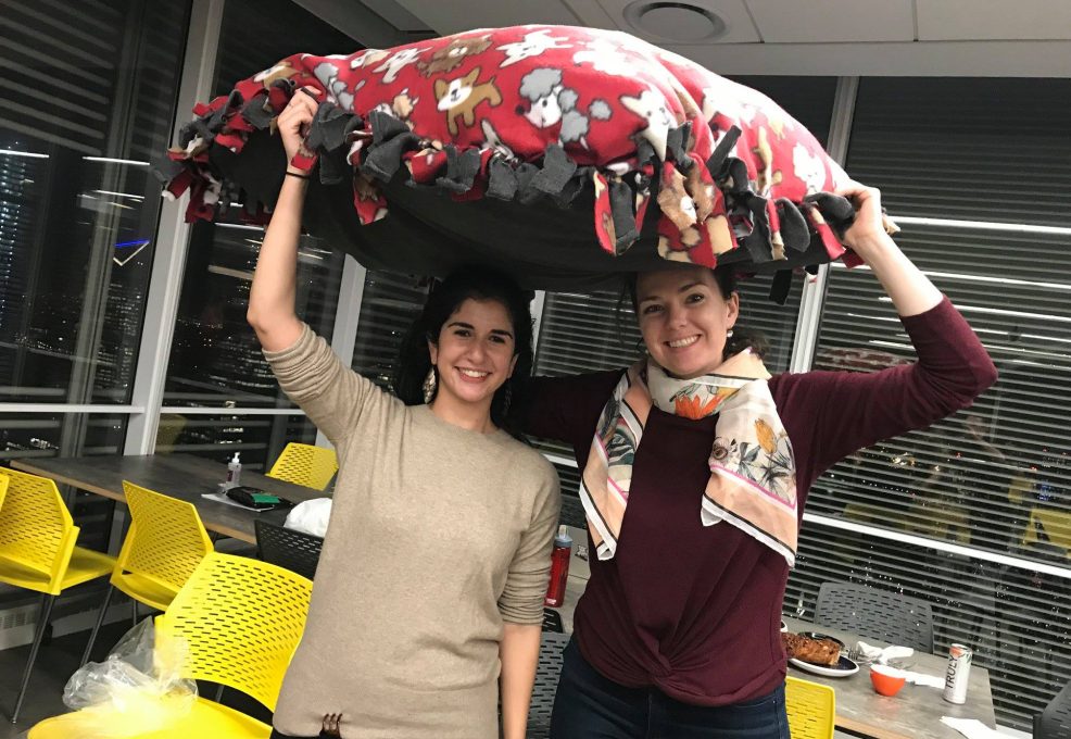 Two smiling participants hold up a large, colorful dog bed they created during the Paws For A Cause team building event, contributing to local animal shelters.