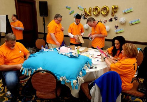 A team dressed in matching orange shirts focuses on crafting a large dog bed and other items during the Paws For A Cause team building event, contributing to local animal shelters.