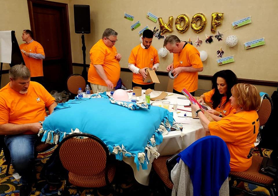 A team dressed in matching orange shirts focuses on crafting a large dog bed and other items during the Paws For A Cause team building event, contributing to local animal shelters.