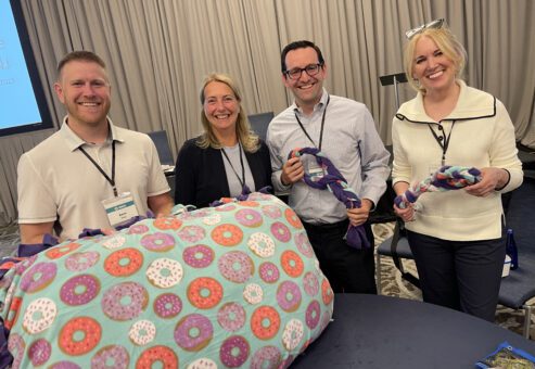 A group of participants from the Paws For A Cause team building event proudly display the dog beds and toys they created, ready to be donated to local animal shelters, with a beautiful waterfront backdrop.