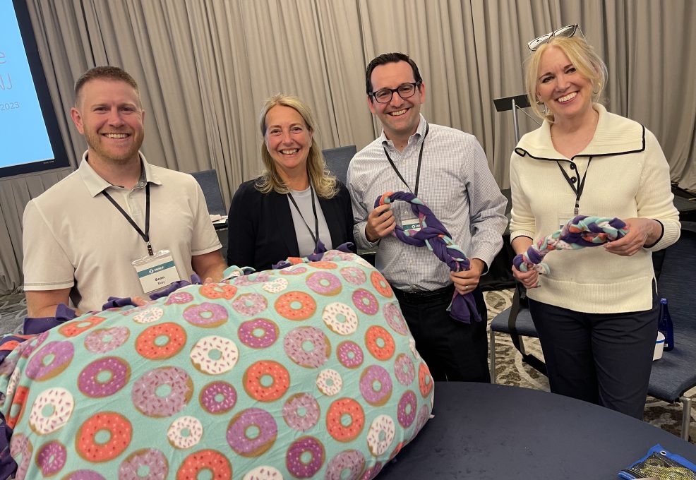 A group of participants from the Paws For A Cause team building event proudly display the dog beds and toys they created, ready to be donated to local animal shelters, with a beautiful waterfront backdrop.
