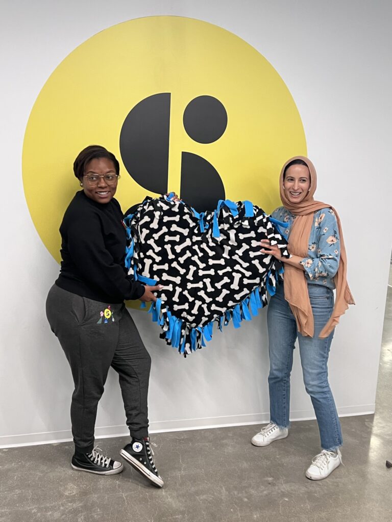 Two participants proudly display a heart-shaped dog bed they created during the Paws For A Cause team building event, supporting local animal shelters with hand-crafted donations.