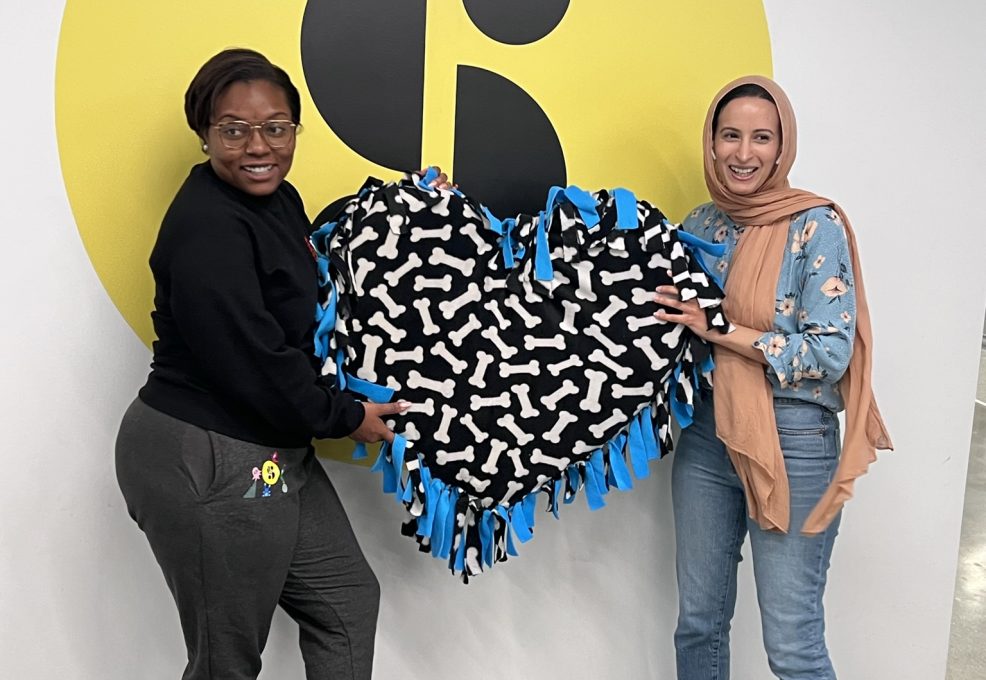 Two participants proudly display a heart-shaped dog bed they created during the Paws For A Cause team building event, supporting local animal shelters with hand-crafted donations.
