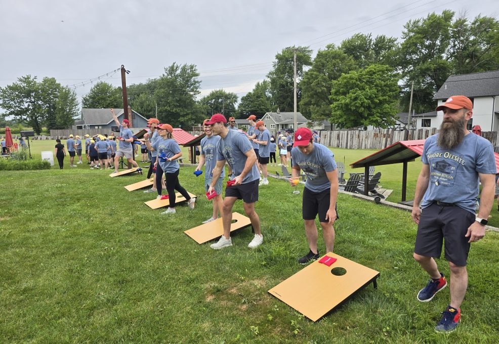 The image shows a group of participants engaged in an outdoor team building activity, specifically playing a game of cornhole. The game involves tossing bean bags into the hole on a raised platform.