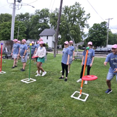 The image shows a group of participants engaged in a team building activity outdoors. The participants are lined up in rows on a grassy field. The activity involves tossing rings or discs onto poles with colored targets. The setup suggests a game that requires accuracy and coordination, promoting teamwork and friendly competition. thumbnail