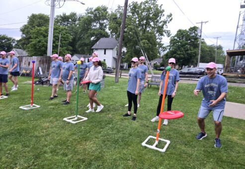 The image shows a group of participants engaged in a team building activity outdoors. The participants are lined up in rows on a grassy field. The activity involves tossing rings or discs onto poles with colored targets. The setup suggests a game that requires accuracy and coordination, promoting teamwork and friendly competition.