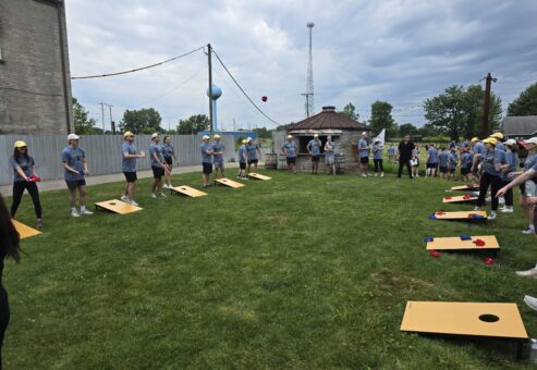 The image shows a group of people participating in an outdoor team building activity. The participants are engaged in a game of cornhole. They are lined up in pairs, tossing bean bags towards wooden boards with holes cut into them.