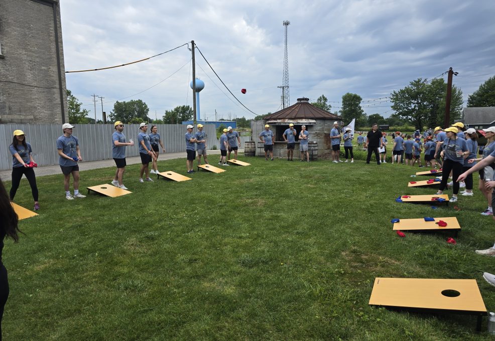 The image shows a group of people participating in an outdoor team building activity. The participants are engaged in a game of cornhole. They are lined up in pairs, tossing bean bags towards wooden boards with holes cut into them.