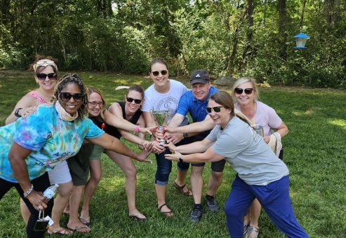A group of teammates smiling and celebrating with a trophy at the Ultimate Tailgate Challenge, posing in the outdoors with classic tailgating games in the background.