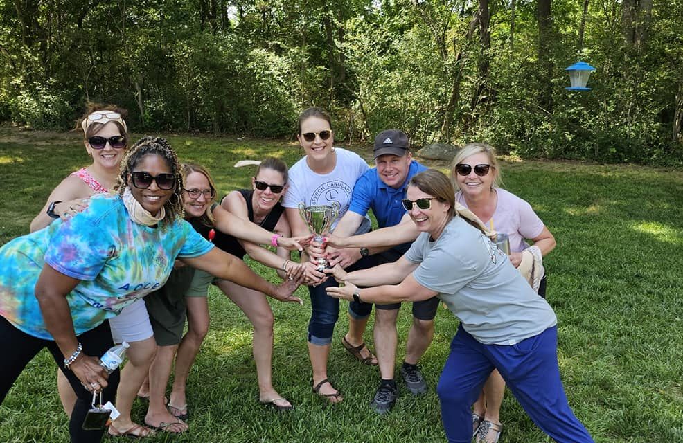 A group of teammates smiling and celebrating with a trophy at the Ultimate Tailgate Challenge, posing in the outdoors with classic tailgating games in the background.