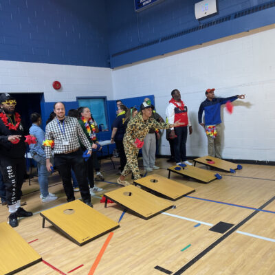 The image captures a group of participants engaging in a game of cornhole during an indoor team building event. The participants are standing behind cornhole boards, taking turns tossing bean bags toward the target. thumbnail