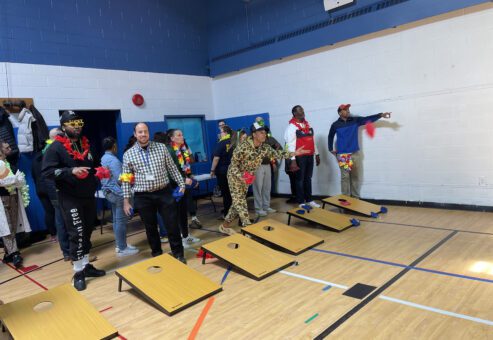 The image captures a group of participants engaging in a game of cornhole during an indoor team building event. The participants are standing behind cornhole boards, taking turns tossing bean bags toward the target.