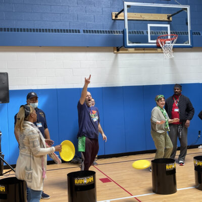 There's a group of people inside a gymnasium engaged in a team building activity. One participant appears to be enthusiastically celebrating or signaling success, adding a lively and energetic atmosphere to the scene. thumbnail