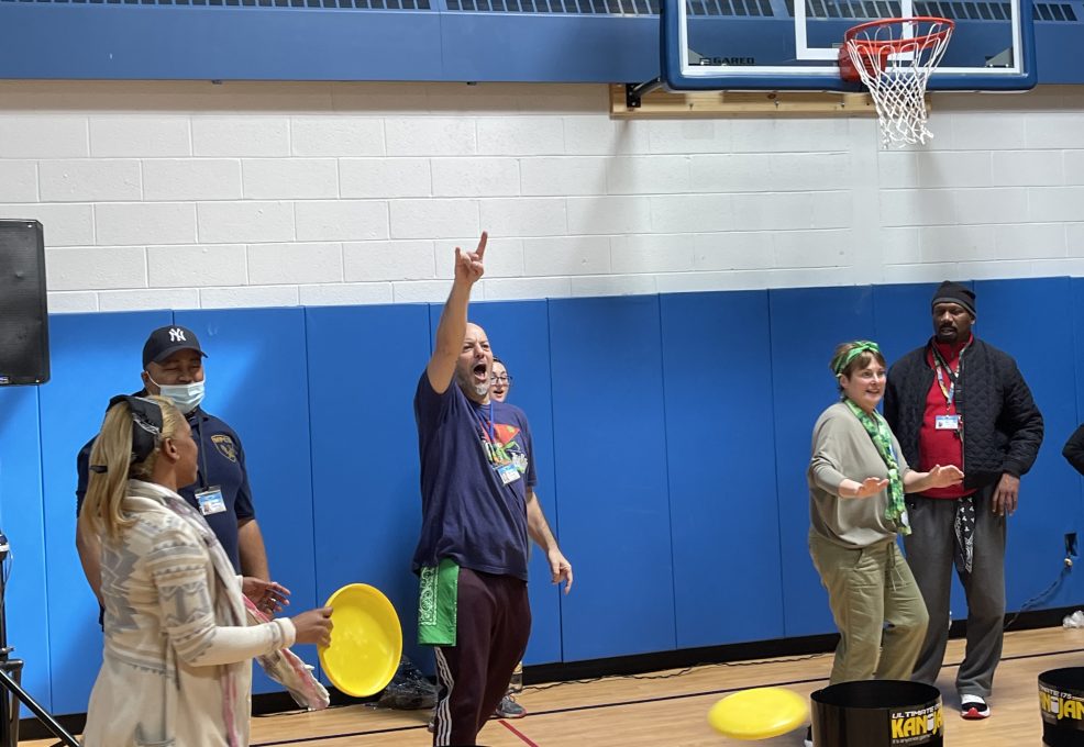 There's a group of people inside a gymnasium engaged in a team building activity. One participant appears to be enthusiastically celebrating or signaling success, adding a lively and energetic atmosphere to the scene.