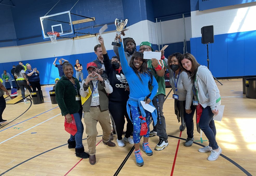 The image shows a group of participants celebrating a victory. The team is gathered in a gymnasium, smiling broadly and holding a trophy, signifying their success. Some members are holding certificates or awards, adding to the celebratory atmosphere. The group appears diverse, with people wearing casual attire, and some are wearing costumes or props, enhancing the fun, playful nature of the event. The joy and camaraderie evident in this image highlight the positive outcomes of team building activities, fostering a sense of unity and accomplishment among participants.