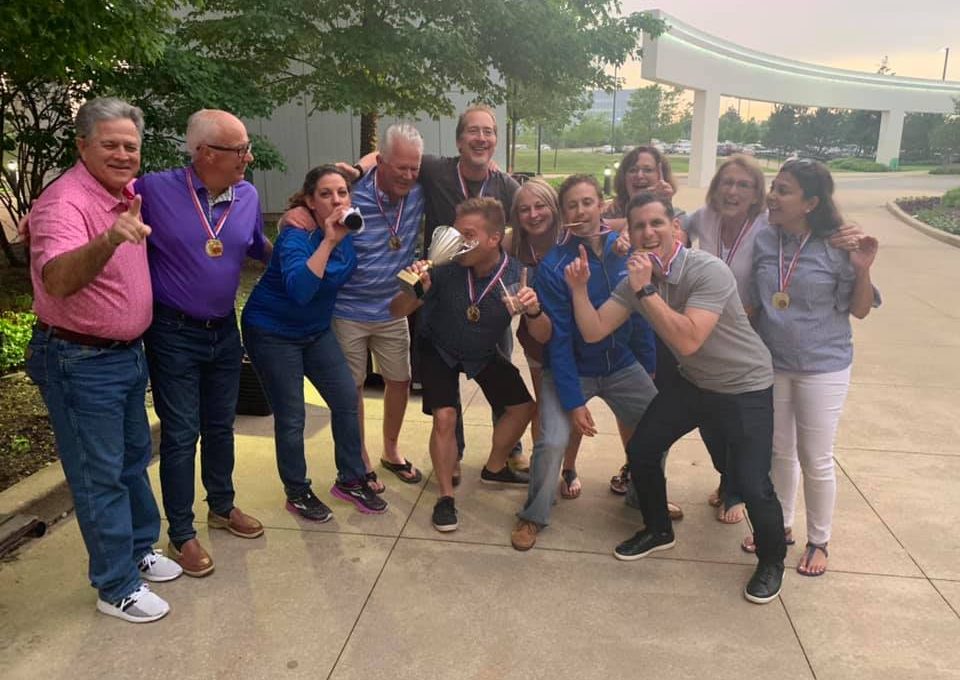 A group of smiling adults posing together outdoors, celebrating their victory at a team building event. They are wearing medals around their necks and holding a trophy, displaying a joyful and triumphant spirit. Some team members are playfully gesturing or mimicking drinking from the trophy, showcasing their camaraderie and enthusiasm.