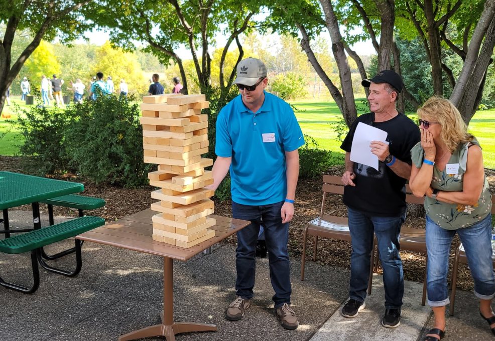 The image shows a group of participants engaged in a giant Jenga game. The participants are focused on the game, watching closely as one person carefully removes a block from the tall, precariously stacked tower.