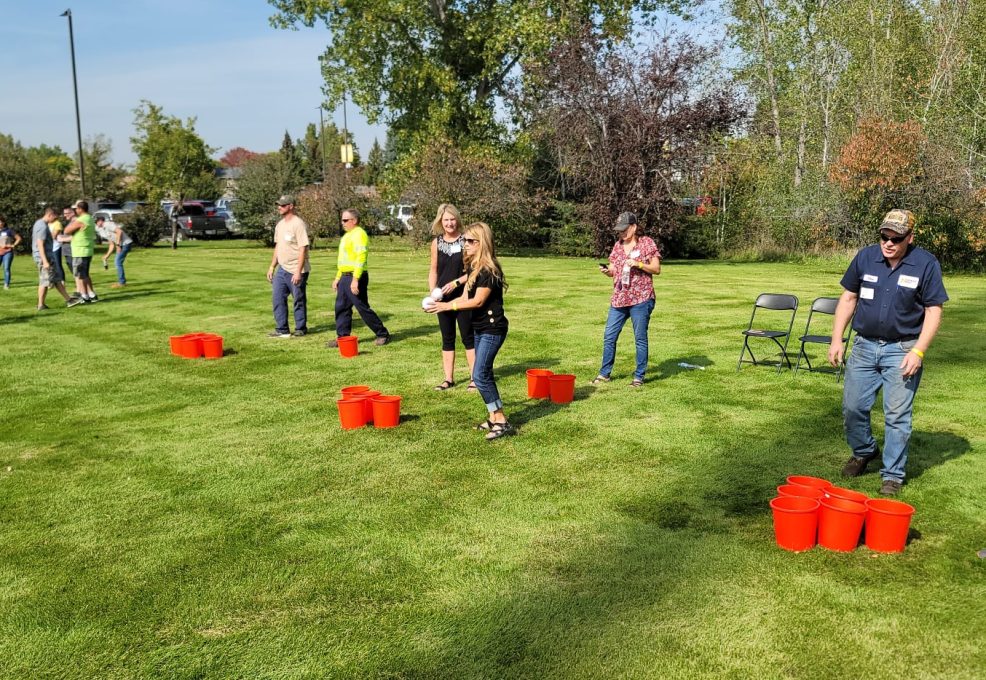 Adults participating in an outdoor team building activity on a sunny day. They are standing on a grassy field, spaced apart, taking part in the Ultimate Tailgate Challenge.