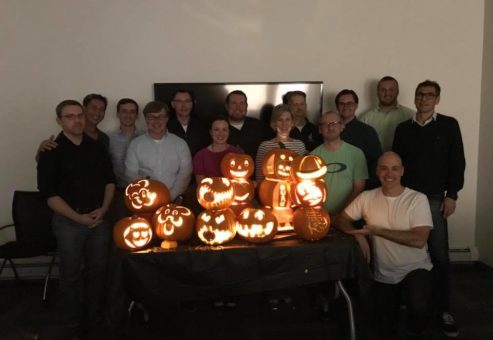 A group of team members proudly display their lit jack-o'-lanterns after a pumpkin carving team building event. The glowing pumpkins feature a variety of creative and fun designs, and the group stands smiling together in a dimly lit room, celebrating their achievements.