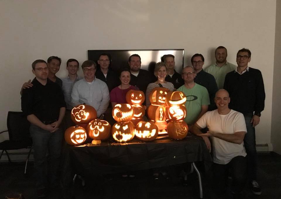 A group of team members proudly display their lit jack-o'-lanterns after a pumpkin carving team building event. The glowing pumpkins feature a variety of creative and fun designs, and the group stands smiling together in a dimly lit room, celebrating their achievements.