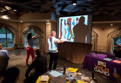 Participants engage in a lively music and dance challenge during the Wild Card Team Building Bingo event, with a vibrant video displayed on the screen.