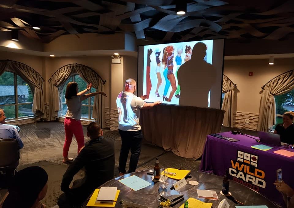 Participants engage in a lively music and dance challenge during the Wild Card Team Building Bingo event, with a vibrant video displayed on the screen.