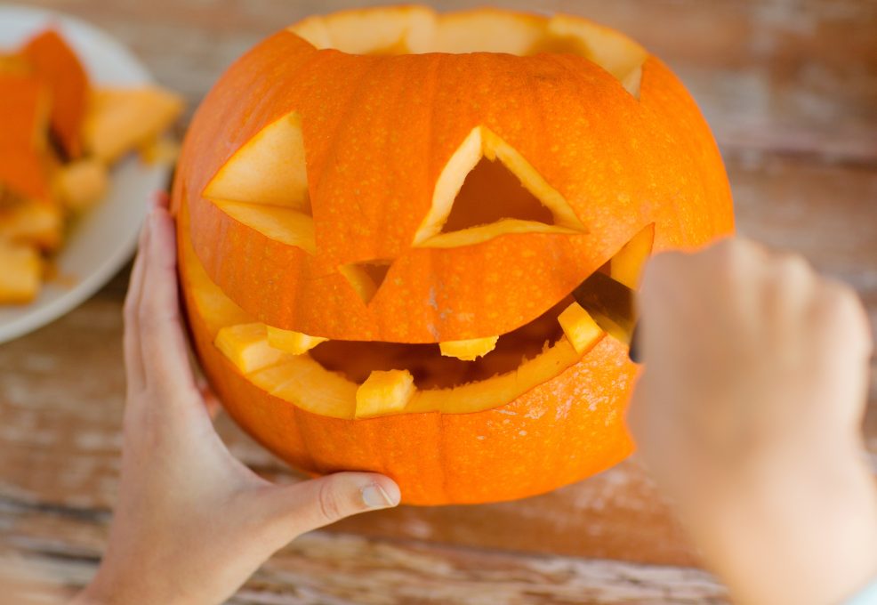 Hands carefully carving a pumpkin, shaping a smiling face with triangular eyes and a wide mouth. Pieces of pumpkin are seen on a plate in the background, showcasing the creative process during a pumpkin carving activity.