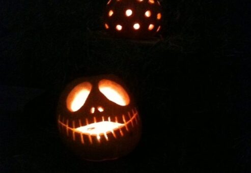 A spooky jack-o'-lantern lit from within, carved with a wide stitched smile and hollow eyes, glowing in the darkness. Another pumpkin with a dotted design is visible in the background, creating an eerie and festive atmosphere during a Halloween-themed team building event.