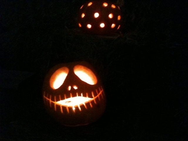A spooky jack-o'-lantern lit from within, carved with a wide stitched smile and hollow eyes, glowing in the darkness. Another pumpkin with a dotted design is visible in the background, creating an eerie and festive atmosphere during a Halloween-themed team building event.