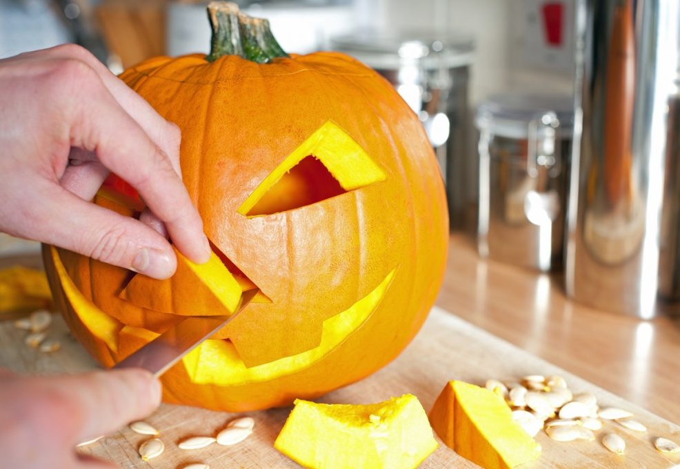 A close-up of hands carving a classic jack-o'-lantern from a pumpkin during a team building event. The partially carved pumpkin features a smiling face, with cut-out pieces and pumpkin seeds scattered on the table, highlighting the creative and hands-on nature of the activity.