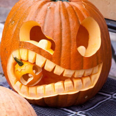 A creatively carved jack-o'-lantern featuring a unique design of a large pumpkin with exaggerated teeth, 'biting' a smaller carved pumpkin. The playful and intricate carving showcases the artistic and humorous approach to pumpkin carving. thumbnail