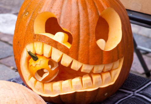 A creatively carved jack-o'-lantern featuring a unique design of a large pumpkin with exaggerated teeth, 'biting' a smaller carved pumpkin. The playful and intricate carving showcases the artistic and humorous approach to pumpkin carving.
