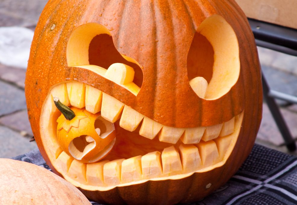 A creatively carved jack-o'-lantern featuring a unique design of a large pumpkin with exaggerated teeth, 'biting' a smaller carved pumpkin. The playful and intricate carving showcases the artistic and humorous approach to pumpkin carving.