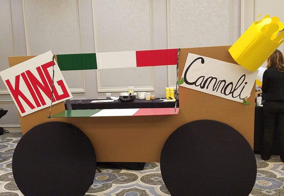 An imaginative 'food truck' display titled 'King Cannoli' is set up as part of a team building event. The mock food truck is constructed with a cardboard facade, and the colors of the Italian flag decorating the upper edge. This creative setup reflects the team's effort to embody an Italian pastry theme for the 'Food Truck Challenge'.