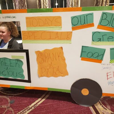 The image showcases a creative and colorful food truck facade crafted for a team building event, focusing on a Greek food theme. The food truck, labeled 