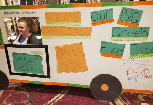 The image showcases a creative and colorful food truck facade crafted for a team building event, focusing on a Greek food theme. The food truck, labeled 