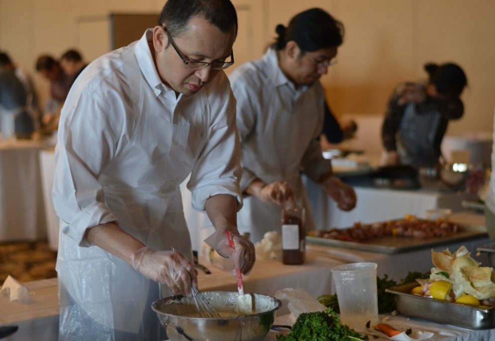A participant at the 'Food Truck Challenge' is preparing a dish. Wearing an apron and gloves, he focuses on mixing ingredients in a large bowl. Behind him, other participants are also engaged in their culinary tasks, surrounded by fresh ingredients