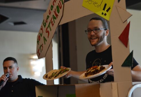 Participants enthusiastically showcase their creative food truck setup during a 'Food Truck Challenge' team building event. A team member stands under a handcrafted food truck façade, proudly presenting plates of tacos.