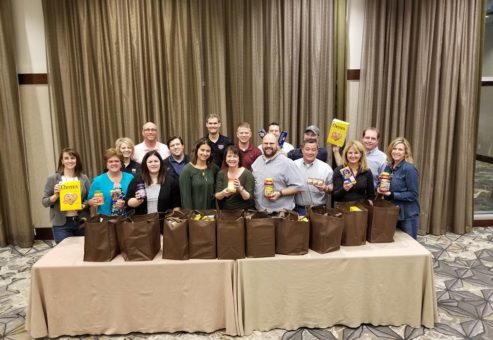A group of participants proudly display bags filled with groceries they packed as part of the Foodbank Showdown team building event, preparing to donate the items to local families in need.