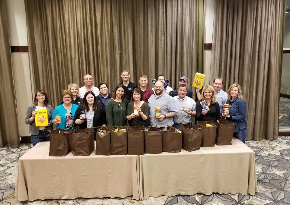 A group of participants proudly display bags filled with groceries they packed as part of the Foodbank Showdown team building event, preparing to donate the items to local families in need.