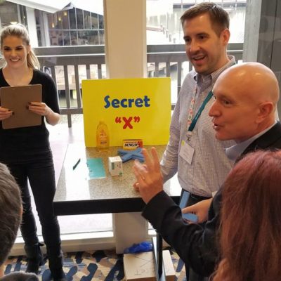 Participants at a Foodbank Showdown team building event engage in grocery-themed game challenges at different stations labeled 'Secret X' and 'Line 'em Up' to win items for donation to families in need. thumbnail