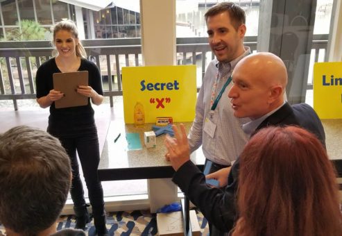 Participants at a Foodbank Showdown team building event engage in grocery-themed game challenges at different stations labeled 'Secret X' and 'Line 'em Up' to win items for donation to families in need.