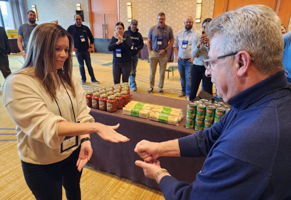 Two participants engage in a rock-paper-scissors game during the Foodbank Showdown team building event. A table filled with canned goods and pasta in the background showcases the charitable aspect of the event.
